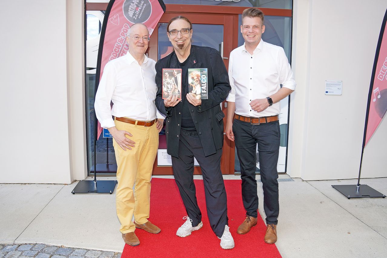 Three men stand on a red carpet in front of a building. The man in the middle holds two books, while the others smile.