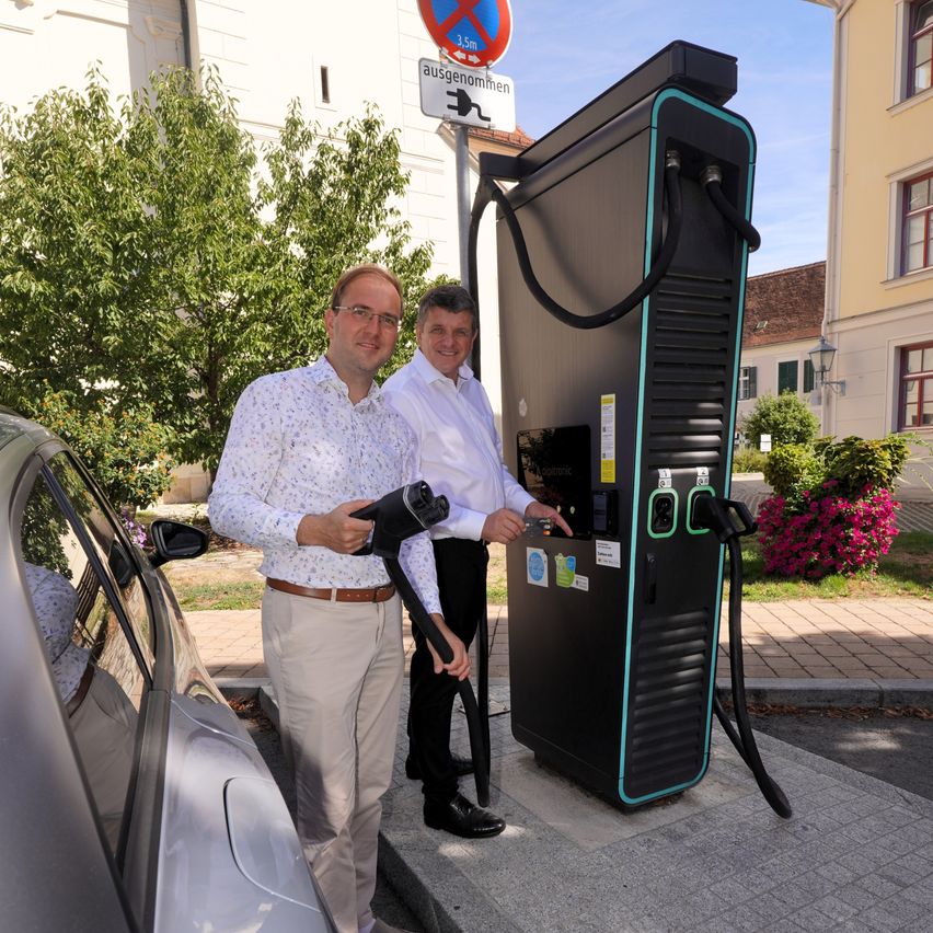 Two men are standing next to a large electric vehicle charging station, with one man holding a charging cable.