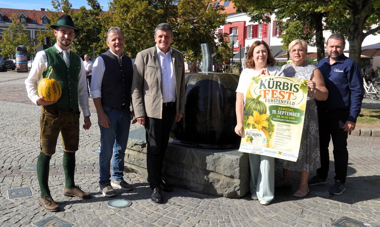 Five people stand in front of a fountain, with one woman holding a poster for the Kürbis Fest.
