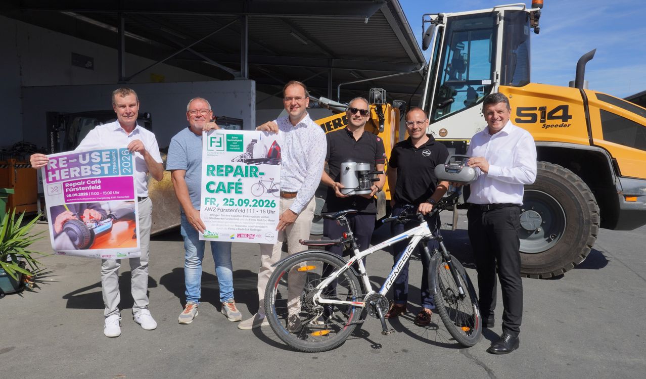 Five men stand in front of a bicycle, one holding a sign that reads 'REPAIR CAFE'. They are posing for a photo. Behind them, a machinery vehicle is parked.