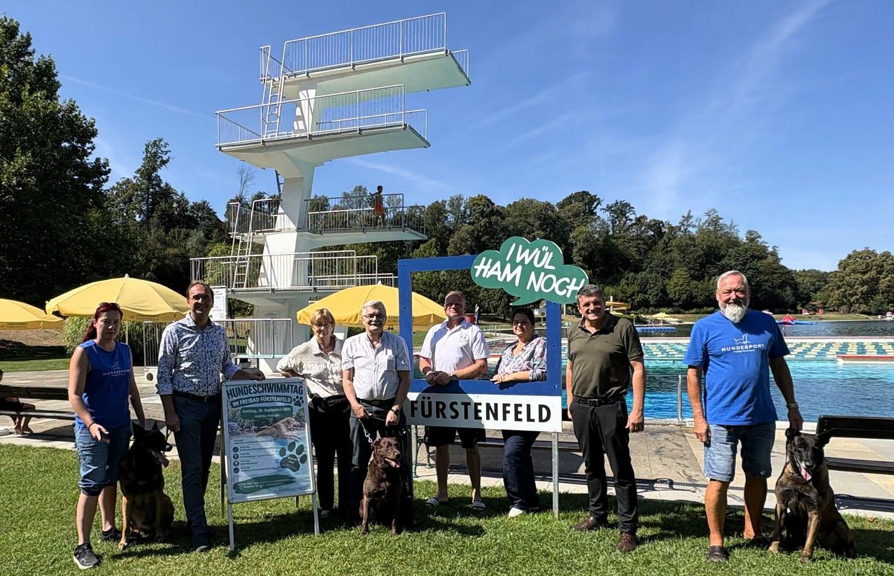 A group of people and a dog stand in front of a sign with a diving tower in the background.