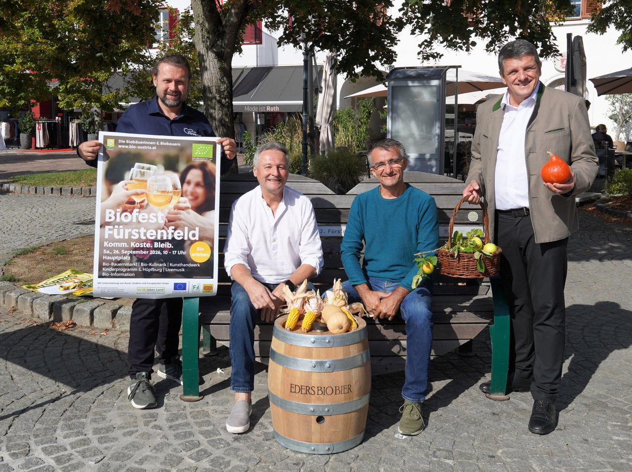 Four men sit on a bench, two holding a poster and two with a barrel of corn and a basket of fruits. They are smiling and posing for a photo.