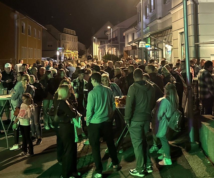 Eine große Menschenmenge versammelt sich auf einer Straße bei Nacht, mit Gebäuden und Straßenlaternen im Hintergrund. Einige tragen Hüte und Turnschuhe, und einige haben Taschen dabei. Ein Tisch mit Essen steht auf dem Bürgersteig.