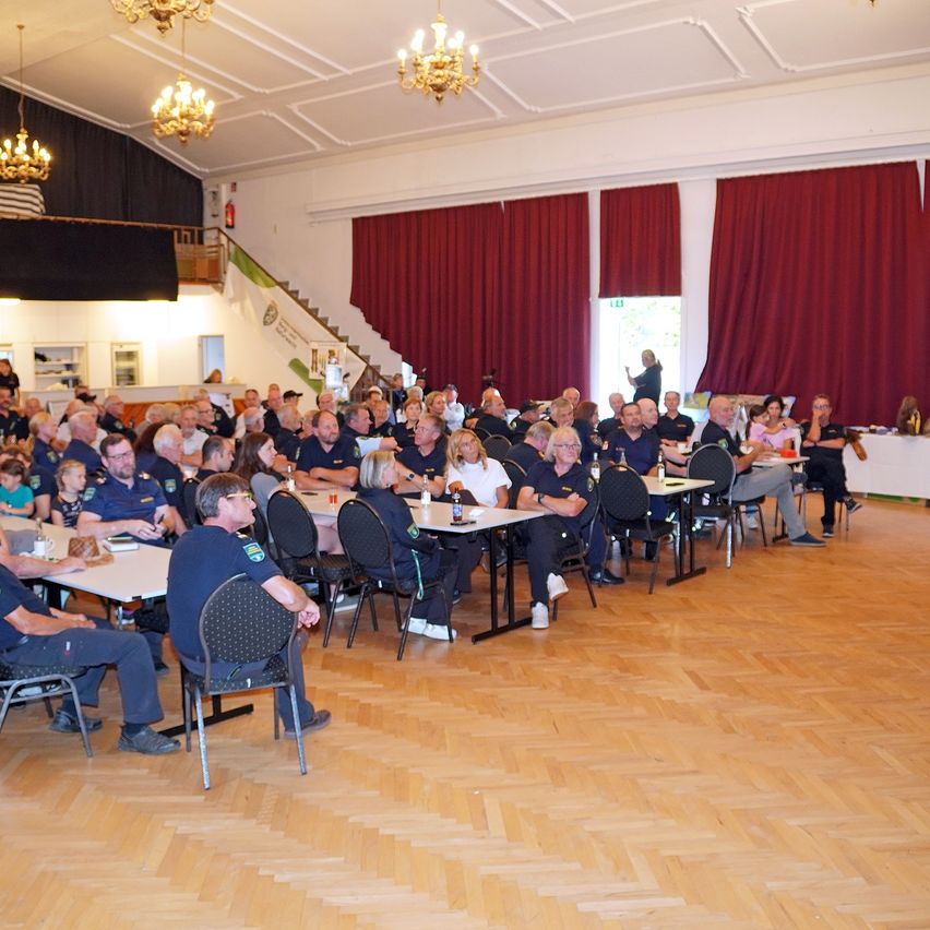 Eine Gruppe von Menschen in Uniform sitzt an Tischen in einem Saal. Einige unterhalten sich, während andere einem Sprecher zuhören. Der Raum hat einen Holzboden und rote Vorhänge.