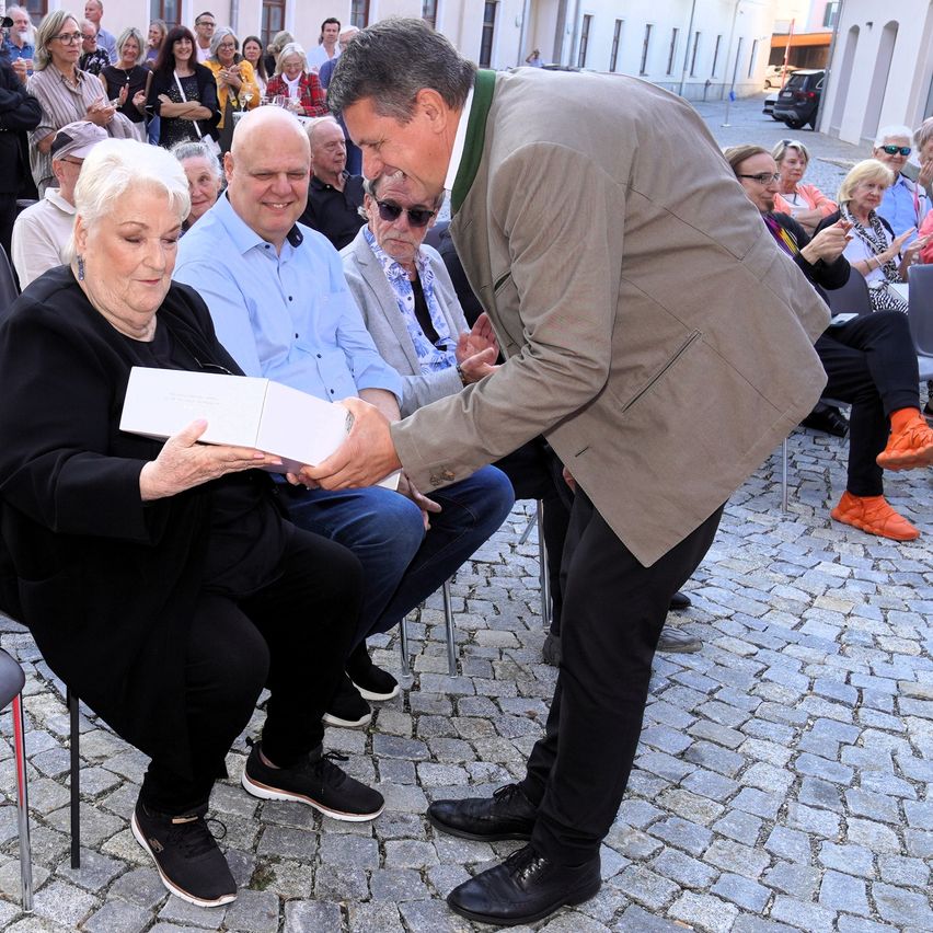 Ein Mann in einem Anzug überreicht einer älteren Frau in einem schwarzen Kleid eine weiße Schachtel während einer Veranstaltung im Freien. Dahinter sitzen mehrere Menschen und schauen zu.