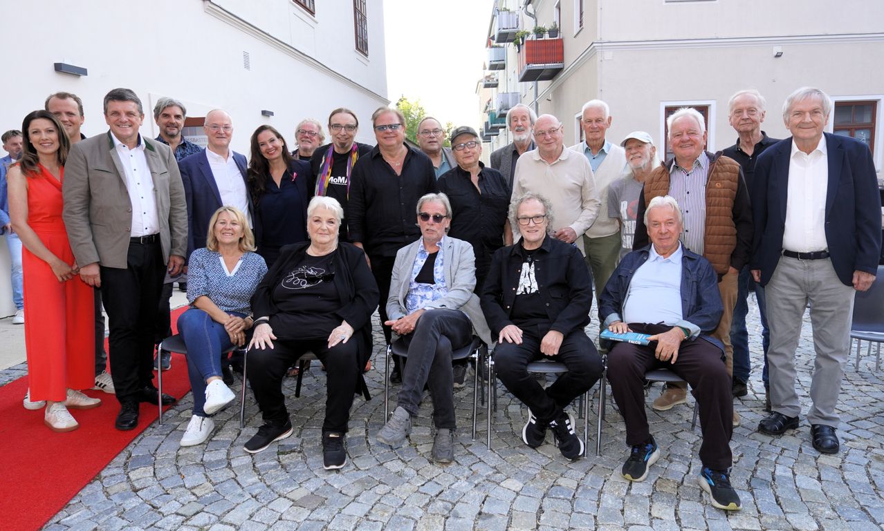Eine Gruppe von Menschen, sowohl Männer als auch Frauen, sitzen auf Stühlen in einer Kopfsteinpflasterstraße und posieren für ein Foto. Sie tragen Freizeitkleidung und einige tragen eine Brille. Hinter ihnen befinden sich Gebäude mit Balkonen.