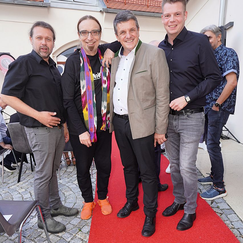 Fünf Männer stehen auf einem roten Teppich und lächeln für ein Foto. Einer trägt einen bunten Schal, ein anderer einen beigen Blazer. Sie sind formell gekleidet. Hinter ihnen stehen zwei weitere Personen, eine mit einer Uhr.
