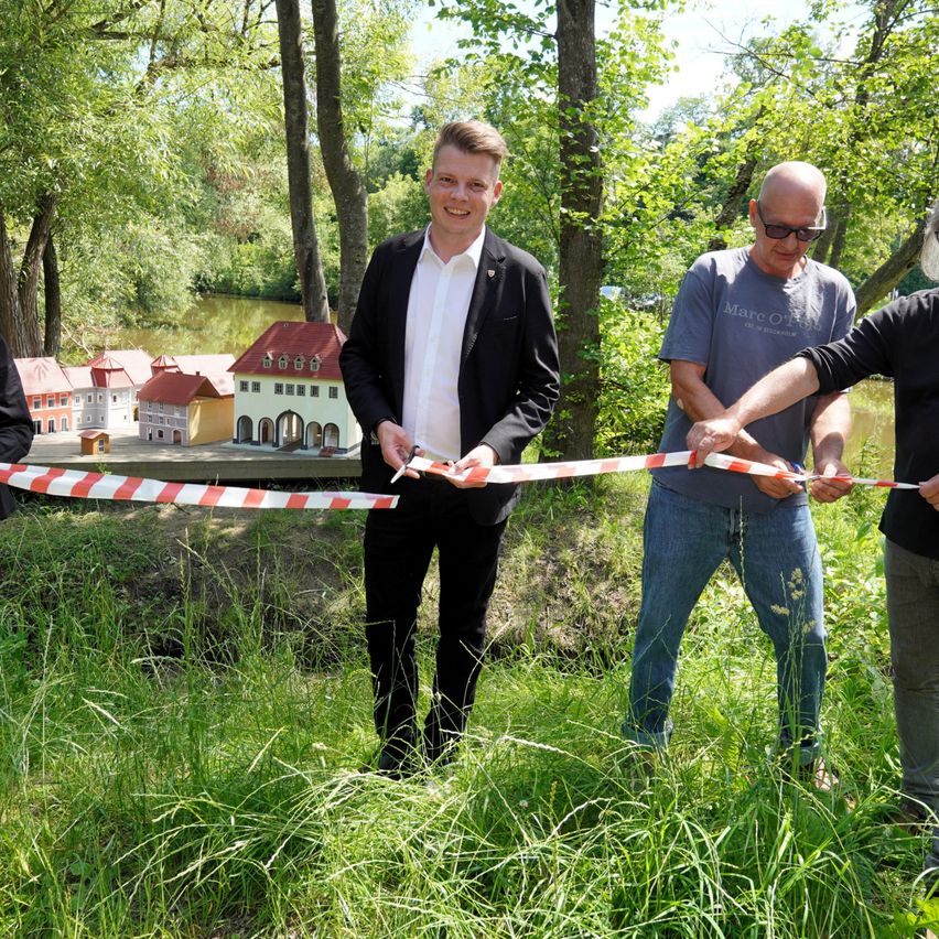 Drei Männer stehen in einem Grasbereich und schneiden ein Band. Dahinter befinden sich Miniaturgebäude und ein See. Der Mann in der Mitte lächelt.