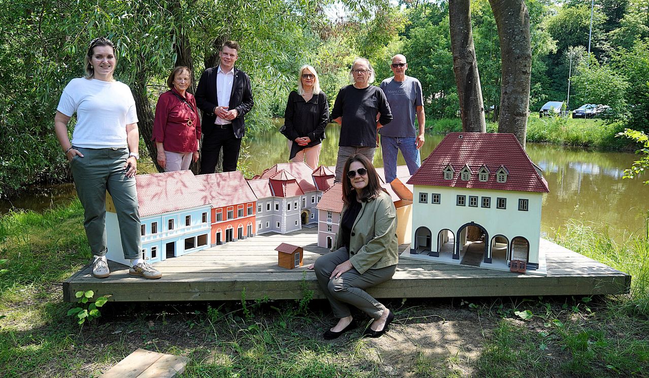 Eine Gruppe von Menschen steht hinter einem Modelldorf, mit einer Frau davor sitzend. Der Hintergrund ist ein Teich mit Bäumen.