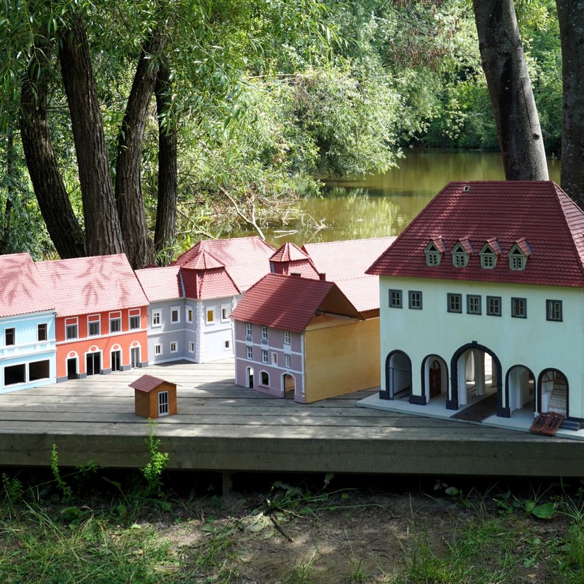 Miniaturmodelle von Gebäuden sind auf einer hölzernen Plattform in der Nähe eines Flusses ausgestellt, umgeben von üppigem Grün und Bäumen.
