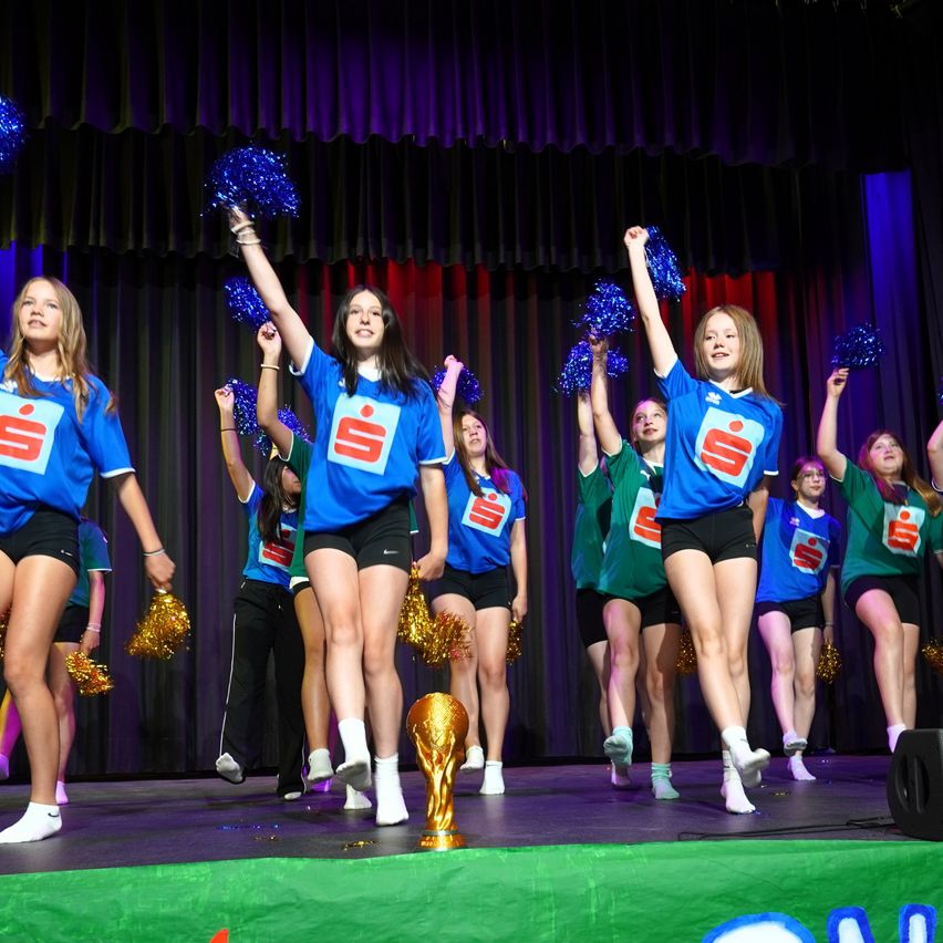 Eine Gruppe von Cheerleadern in blauen Shirts mit rotem 'S' performt auf der Bühne mit einem goldenen Pokal.