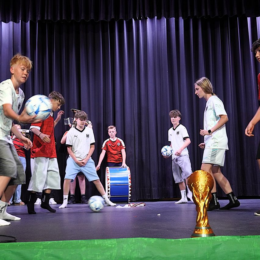 Eine Gruppe junger Jungen spielt Fußball auf einer Bühne. Einige halten Fußballbälle und andere treten sie. Ein Pokal ist vor ihnen aufgestellt.
