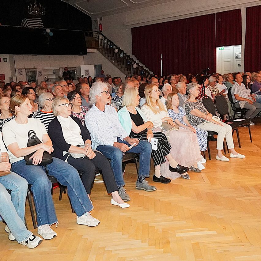 Ein voll besetzter Saal mit Besuchern in Reihen. Einige tragen Brillen, und einige halten Papiere. Der Raum hat Holzböden und rote Vorhänge.