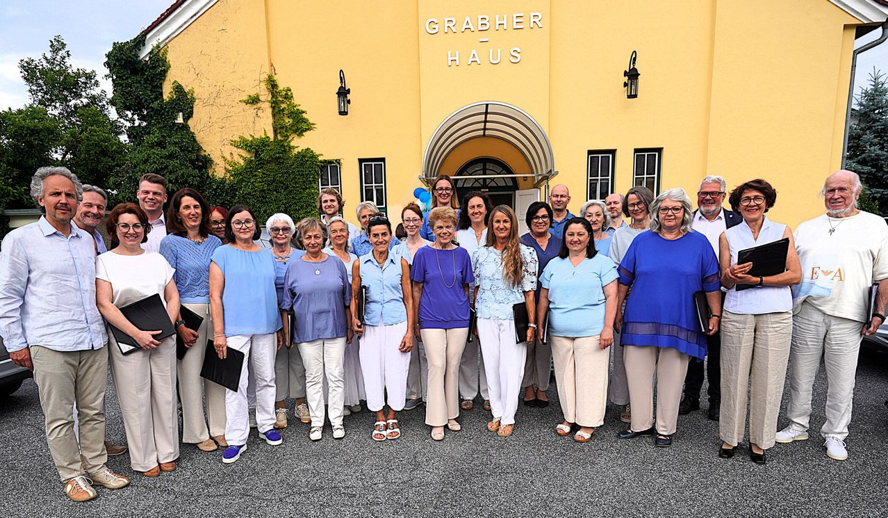 Eine Gruppe von Menschen, hauptsächlich Frauen, posiert für ein Foto vor dem Grabher Haus. Sie tragen aufeinander abgestimmte Outfits und einige halten Bücher. Das Gebäude hat gelbe Wände und ein Schild, das 'Grabher Haus' steht.