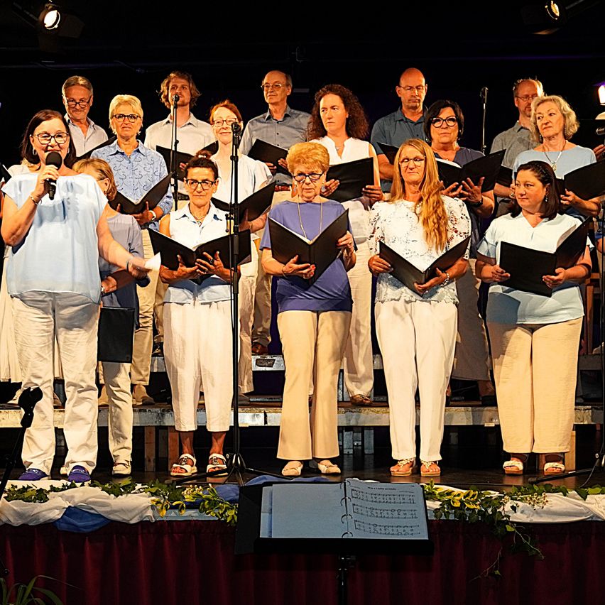 Ein Chor tritt auf der Bühne auf, hält Notenblätter in der Hand, mit einem Dirigenten vorne, unter Scheinwerfern.
