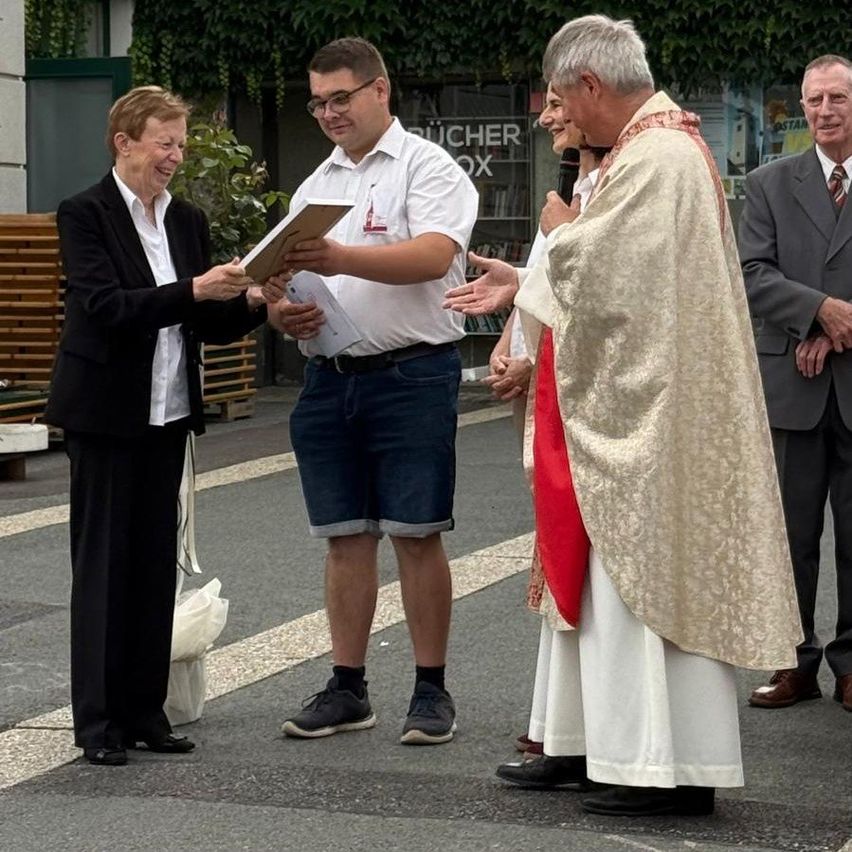 Ein Priester überreicht einer Frau ein Dokument während einer Zeremonie auf einer Stadtstraße. Hinter ihnen beobachten zwei Männer.