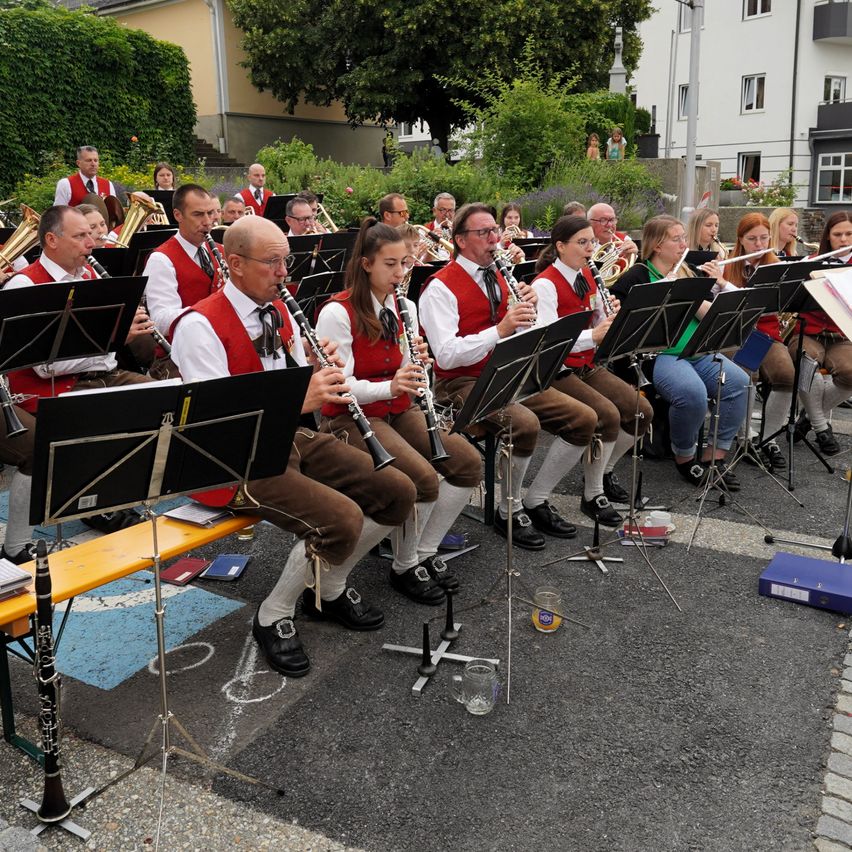 Eine Band übt an einem sonnigen Tag auf einem Stadtplatz, mit Musikern in roten Westen und weißen Socken, die Instrumente wie Klarinetten und Trompeten spielen.