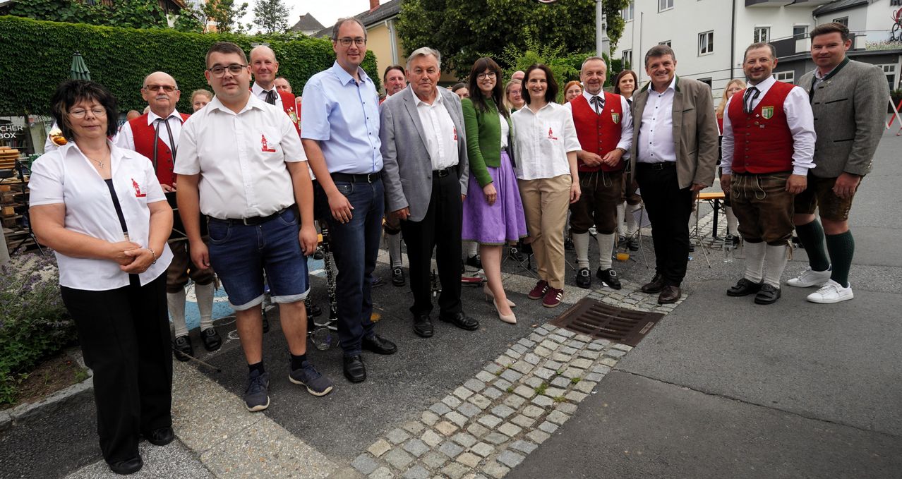 Eine Gruppe von Menschen, einschließlich Männer und Frauen, steht draußen auf einer Kopfsteinpflasterstraße. Sie sind in Freizeit- und traditioneller Kleidung gekleidet. Gebäude und Grünflächen sind im Hintergrund.