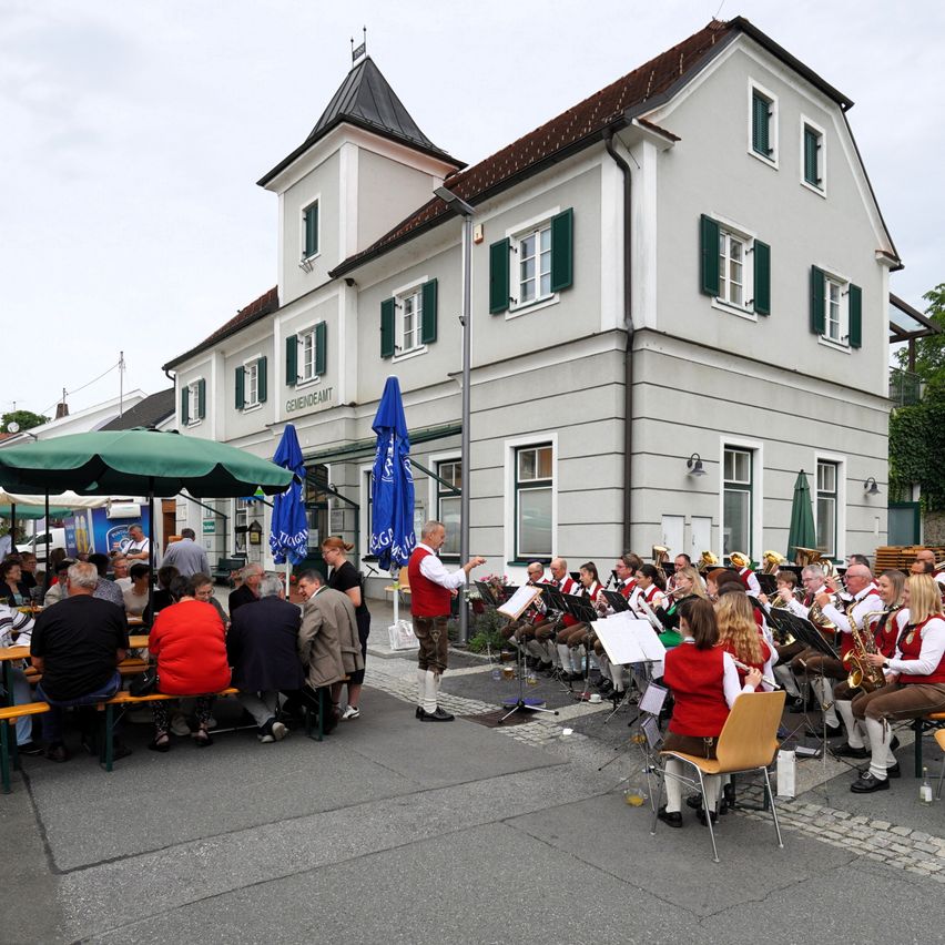Eine Band spielt vor einem großen Gebäude mit vielen Fenstern und einem Turm, während Menschen zuhören. Einige Leute sitzen auf Bänken, andere stehen. Die Bandmitglieder spielen verschiedene Musikinstrumente.