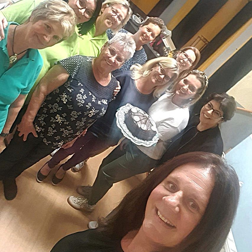 Eine Gruppe von Frauen lächelt und posiert für ein Foto, eine hält einen Kuchen. Sie sind drinnen mit einem Holzboden.