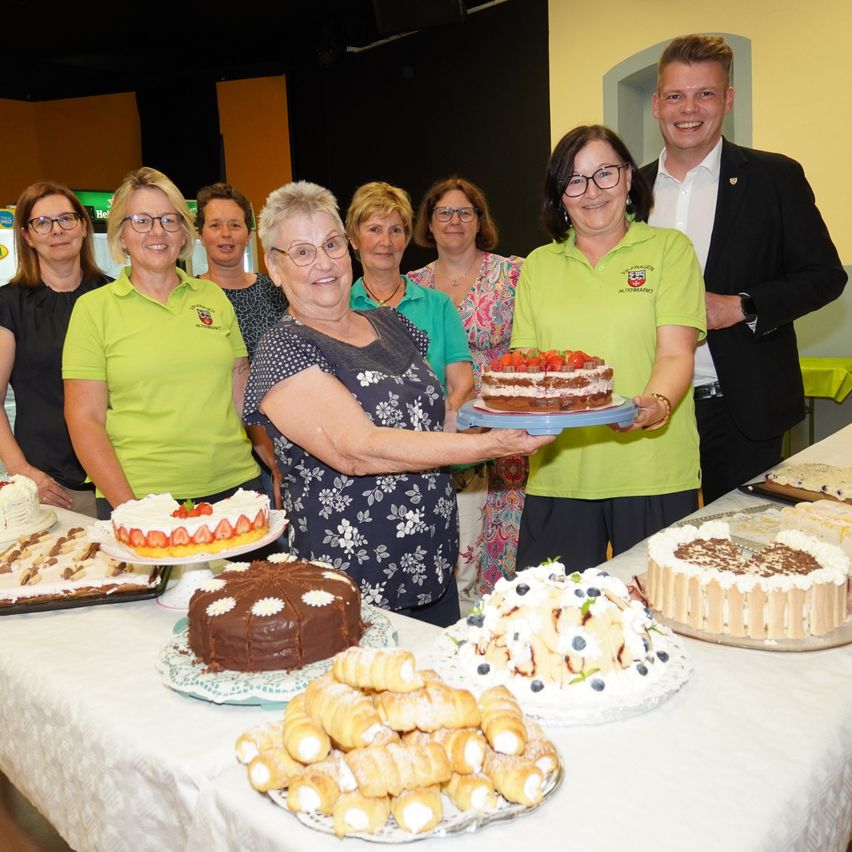 Eine Gruppe von Menschen in grünen Shirts steht hinter einem Tisch voller Kuchen und Gebäck, wobei zwei Frauen eine Platte mit Kuchen halten. Hinter ihnen lächelt ein Mann im Anzug.