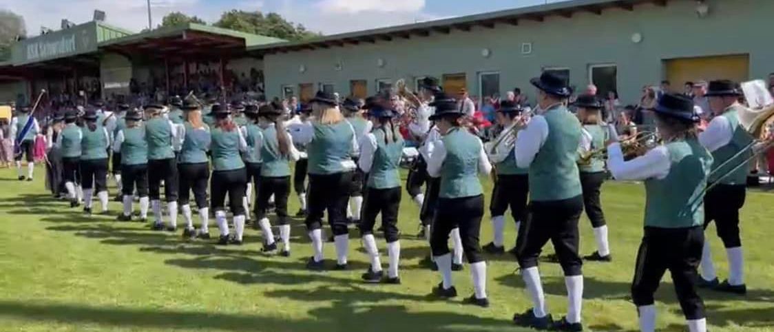 Eine Musikkapelle in traditionellen Uniformen tritt auf einem Rasenfeld auf, während Zuschauer von hinten zusehen. Die Bandmitglieder spielen Trompeten und Posaunen.