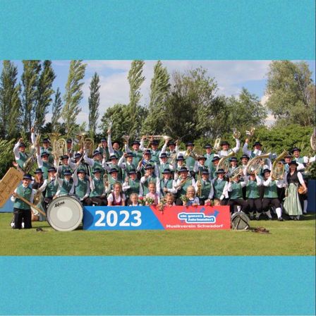 Eine große Gruppe von Musikern in grünen Uniformen, einige mit Hüten, posiert für ein Foto mit Instrumenten und einem Banner, das 2023 sagt.