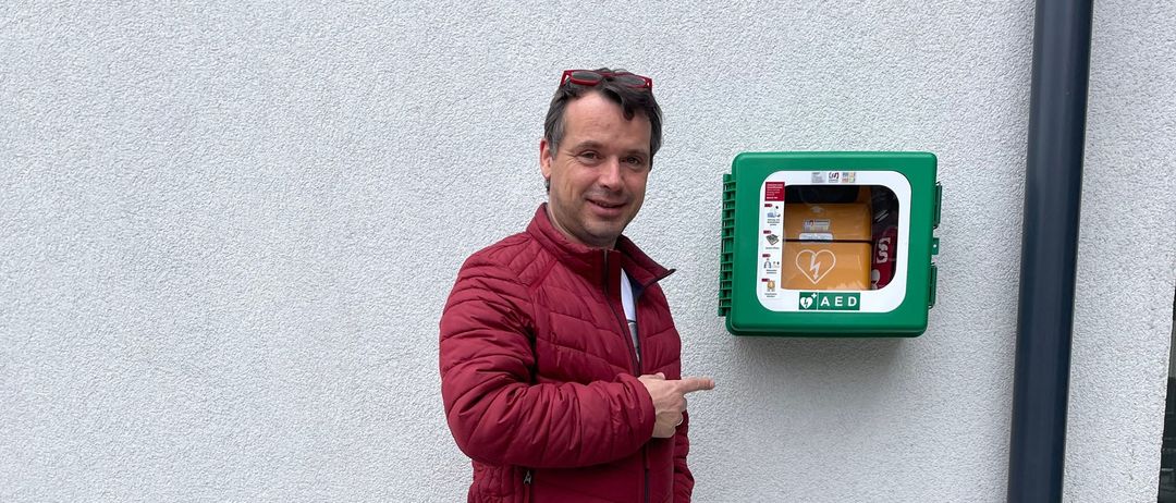 Ein Mann in einem roten Jackett und blauen Jeans steht neben einer weißen Wand und zeigt auf eine grün-weiße AED-Box an der Wand.