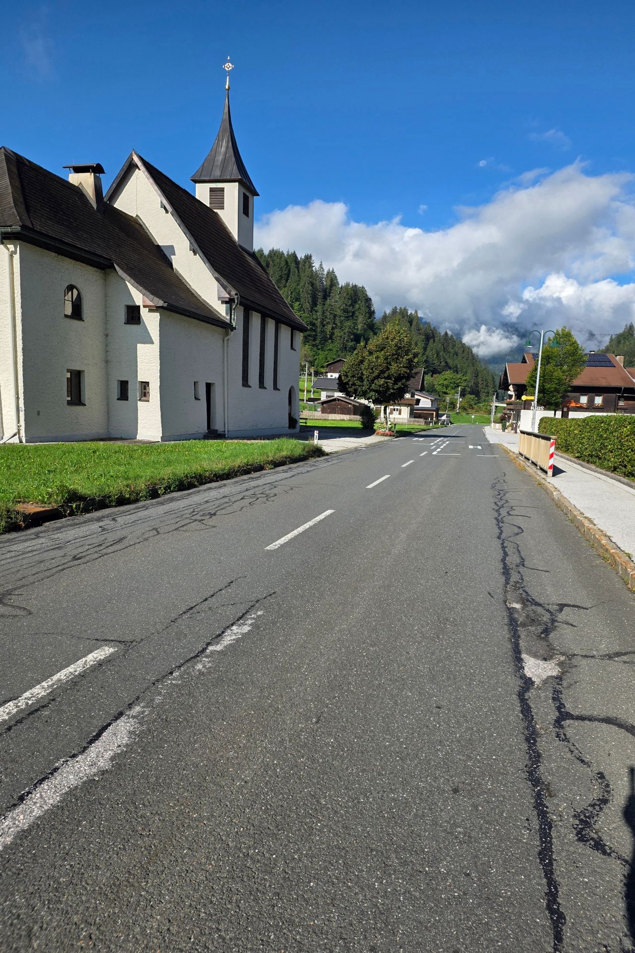 Eine breite, leere Straße mit einer Kirche links und Häusern rechts, unter einem teilweise bewölkten Himmel.