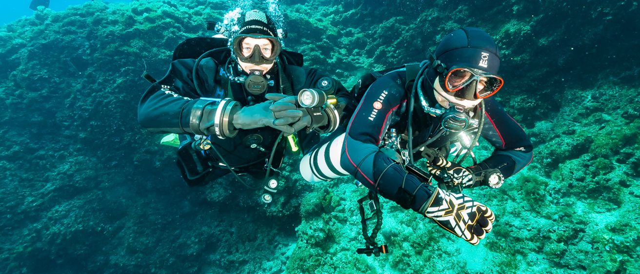 Zwei Taucher sind unter Wasser, umgeben von Korallenriffen. Ein Taucher hält einen Atemregler und ein Manometer. Beide tragen schwarze Helme, Schutzbrillen und Tauchanzüge.