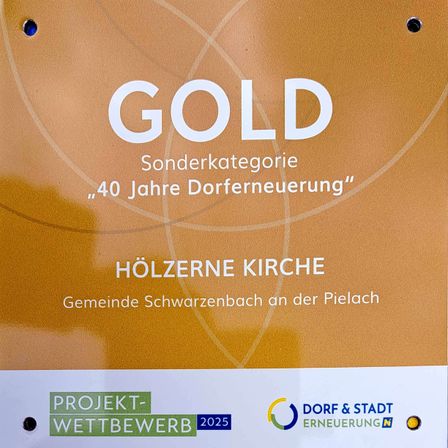 Ein goldenes Schild mit weisser Schrift zeigt 'GOLD', eine Sonderkategorie für '40 Jahre Dorferneuerung'. Darunter steht 'Hölzerne Kirche', gefolgt von 'Gemeinde Schwarzenbach an der Pielach'. Unten befindet sich 'Projekt-Wettbewerb 2025'.