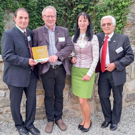Vier Menschen stehen gemeinsam vor einer Steinmauer. Ein Mann hält eine goldene Plakette mit Text. Sie tragen alle formelle Kleidung.