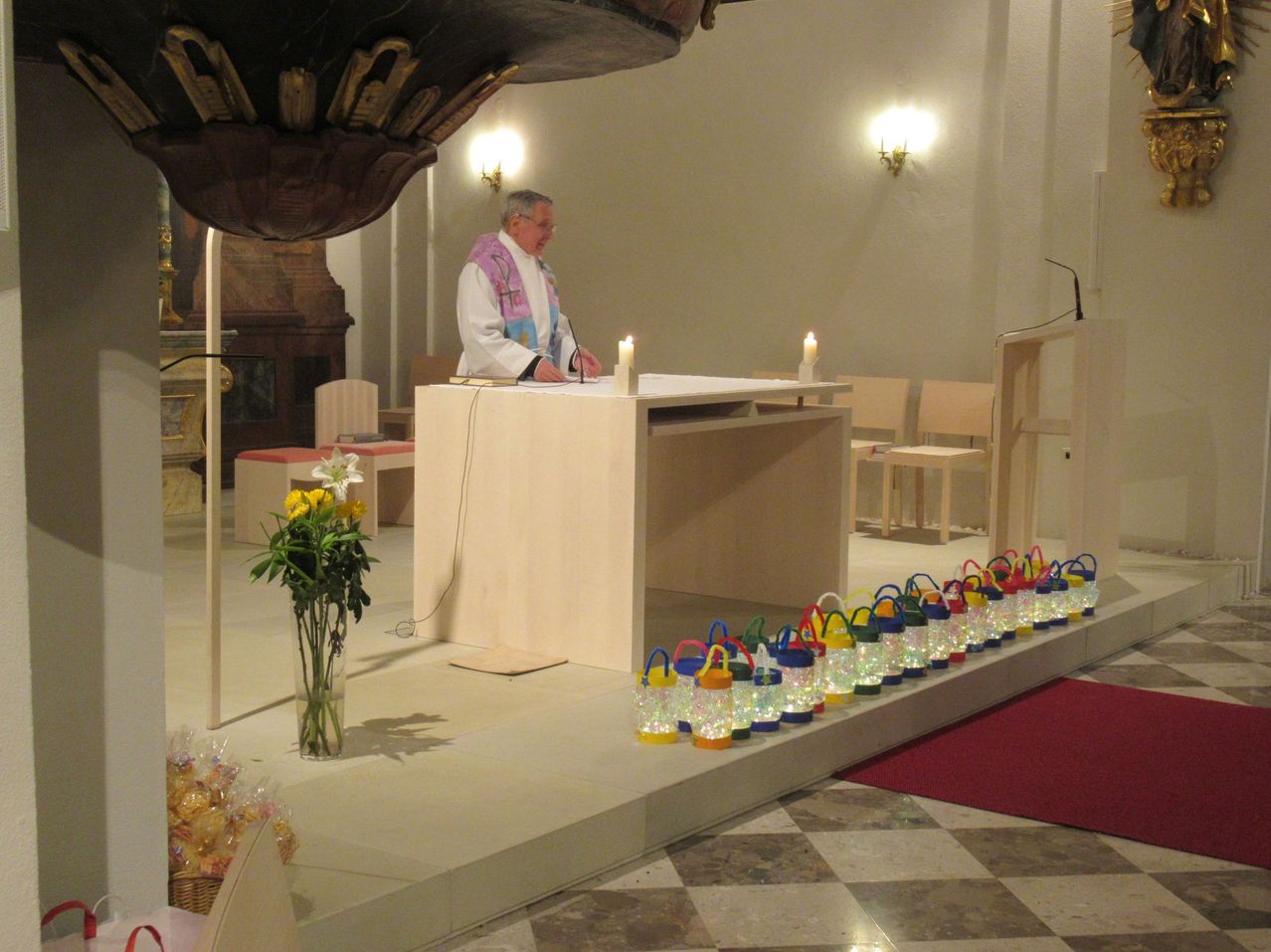 Ein Priester steht an einem Podium in einer Kirche, umgeben von Kerzen und Blumen. Bunte Laternen säumen den Altar.