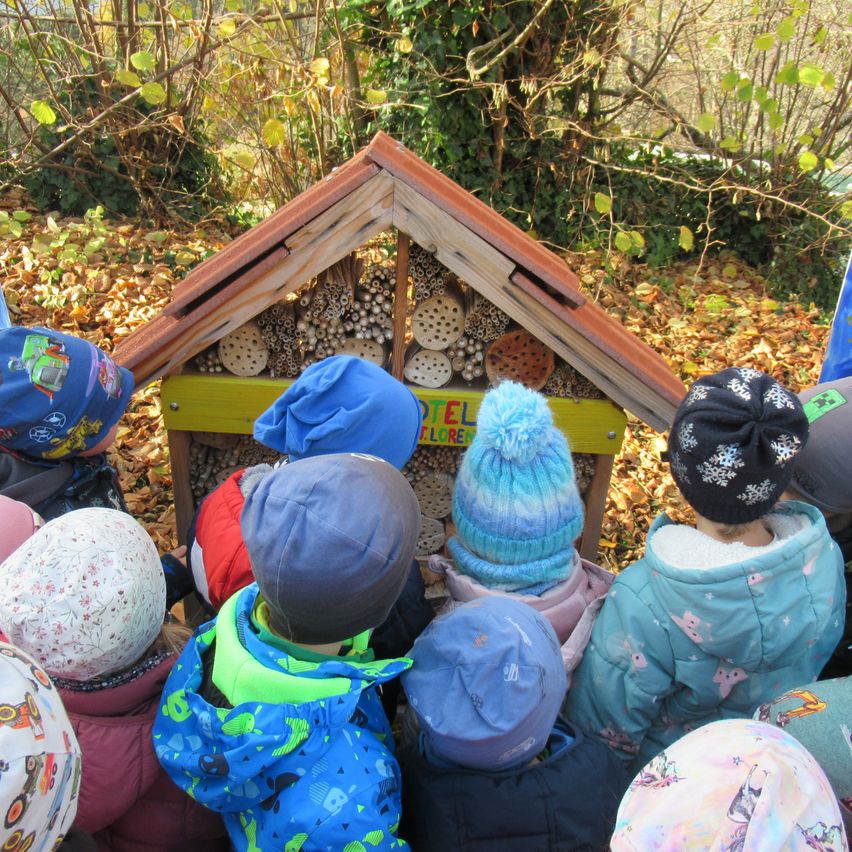 Eine Gruppe von Kindern hat sich um eine kleine Holzstruktur in einem Garten versammelt, möglicherweise ein Bienenhotel, und trägt Winterkleidung und Mützen.