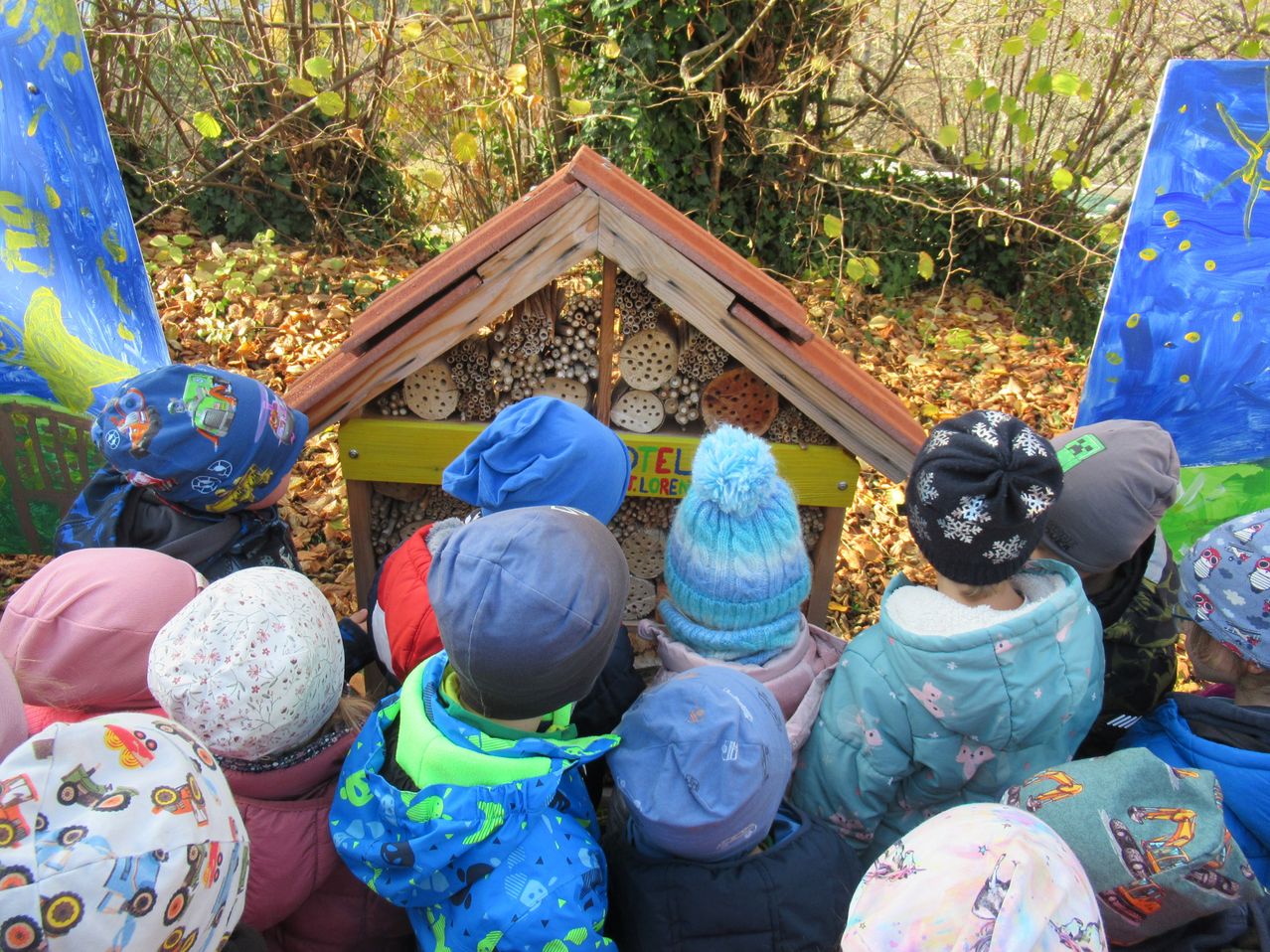 Eine Gruppe von Kindern hat sich um eine kleine Holzstruktur in einem Garten versammelt, möglicherweise ein Bienenhotel, und trägt Winterkleidung und Mützen.