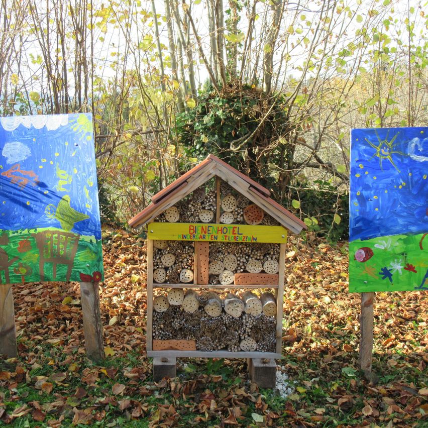 Ein Insektenhotel mit zahlreichen Löchern in einer Waldumgebung, umgeben von zwei bemalten Tafeln. Die Tafeln haben farbenfrohe Kunstwerke, und der Boden ist mit trockenen Blättern bedeckt.