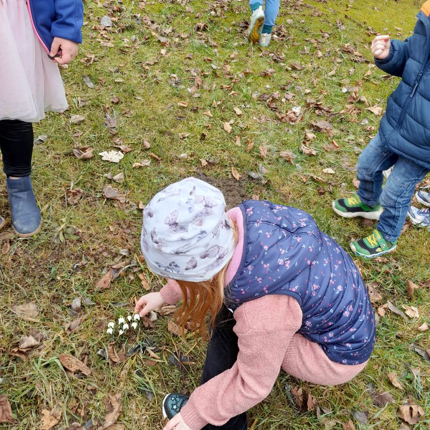 Ein junges Mädchen kniet auf dem Boden und pflückt Blumen. Andere Kinder stehen und gehen in der Nähe. Der Boden ist mit trockenen Blättern bedeckt.