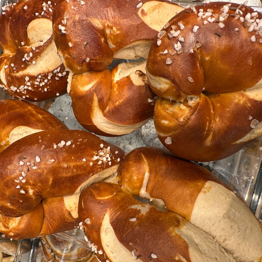 A close-up view of pretzels with a golden-brown crust and sprinkled salt. The pretzels are slightly flattened, and some are folded in half. They are displayed on a metal tray.