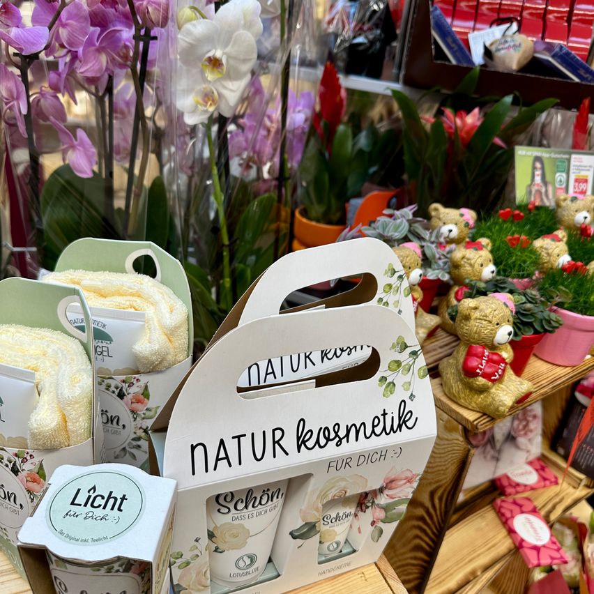 Display of natural cosmetics in a shop with floral background, including a box labeled 'Natur Kosmetike' with a floral design and two small boxes inside. Various plants and flowers are in the background.
