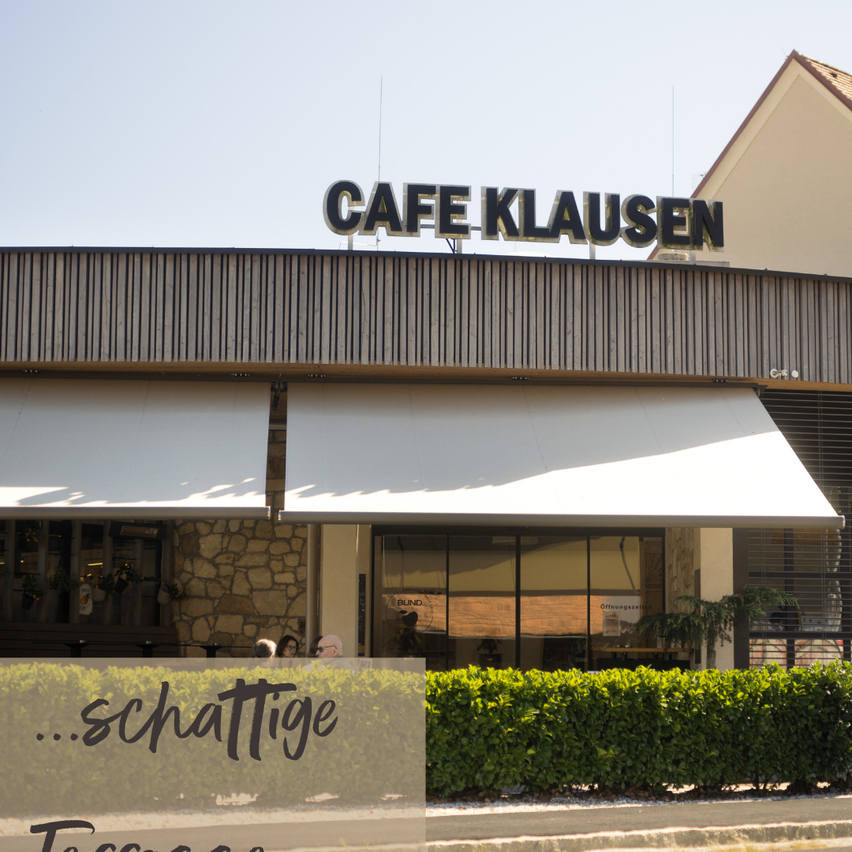 Ein Café namens Cafe Klausen mit einem weißen Vordach und einer Steinmauer-Außenwand. Das Schild ist auf dem Dach montiert.