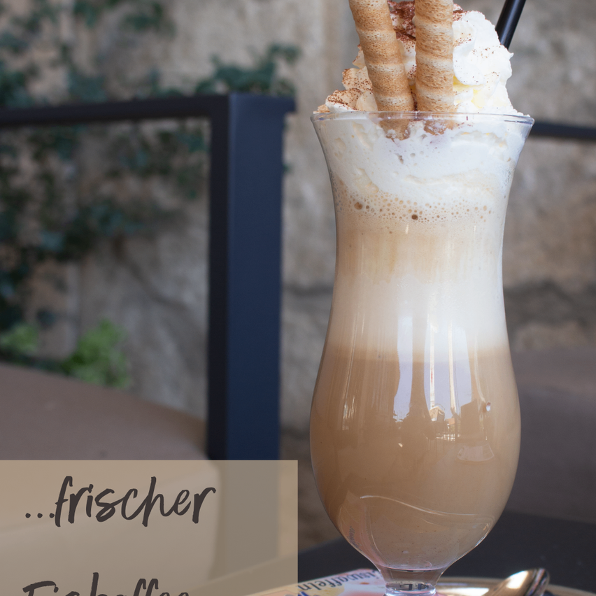Ein hohes Glas mit einem cremigen Kaffeegetränk, gekrönt mit Schlagsahne, Schokoladenstreuseln und zwei Zimtschnecken. Das Getränk ist mit einem schwarzen Strohhalm garniert und steht auf einem Tisch.
