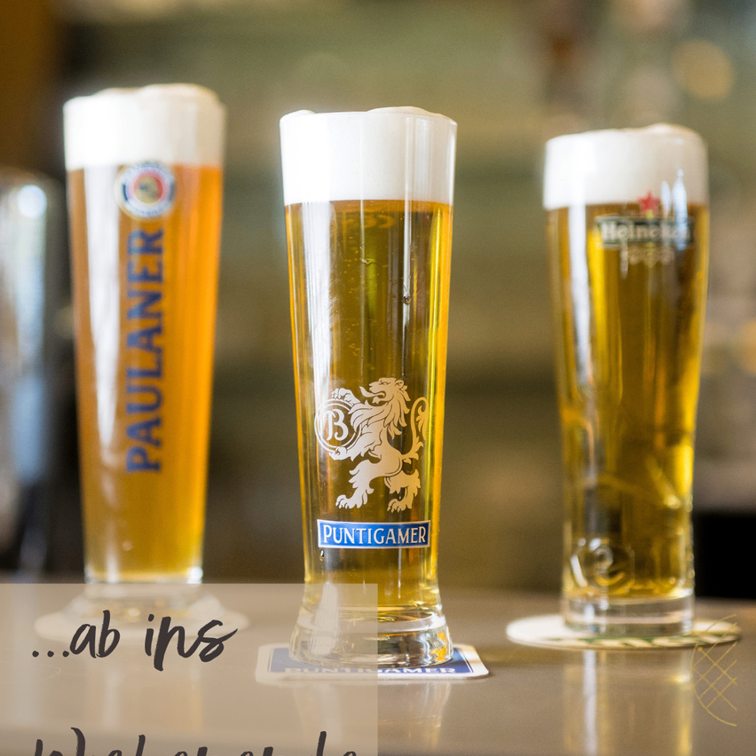 Drei hohe Biergläser auf einer Bar, darunter ein Glas Paulaner, ein Glas Puntigamer mit Löwenlogo und ein Glas Heineken.