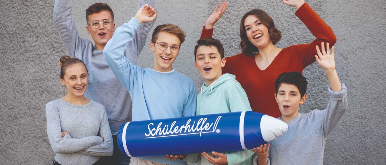 Eine Gruppe von Teenagern posiert für ein Foto mit einem großen blauen aufblasbaren Bleistift von Schillerhilfe. Sie alle haben große Lächeln auf ihren Gesichtern.