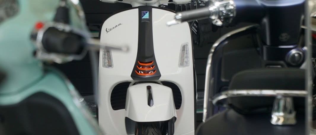 Ein weißer Vespa Roller mit schwarzem Akzent und orangefarbenen Lichtern steht in einer Ausstellung.