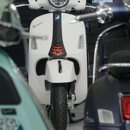 Nahaufnahme eines weißen Vespa Rollers mit schwarzem Sitz und orangefarbenen Akzenten, der in einer Ausstellungshalle geparkt ist.