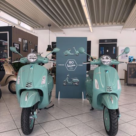 Zwei mintgrüne Vespa-Roller werden in einer Ausstellungshalle präsentiert, hinter ihnen ein blauer Banner, auf dem '80 Jahre für immer ikonisch' steht. Die Roller stehen auf einem gefliesten Boden, und im Hintergrund befinden sich weitere Roller und Gegenstände.