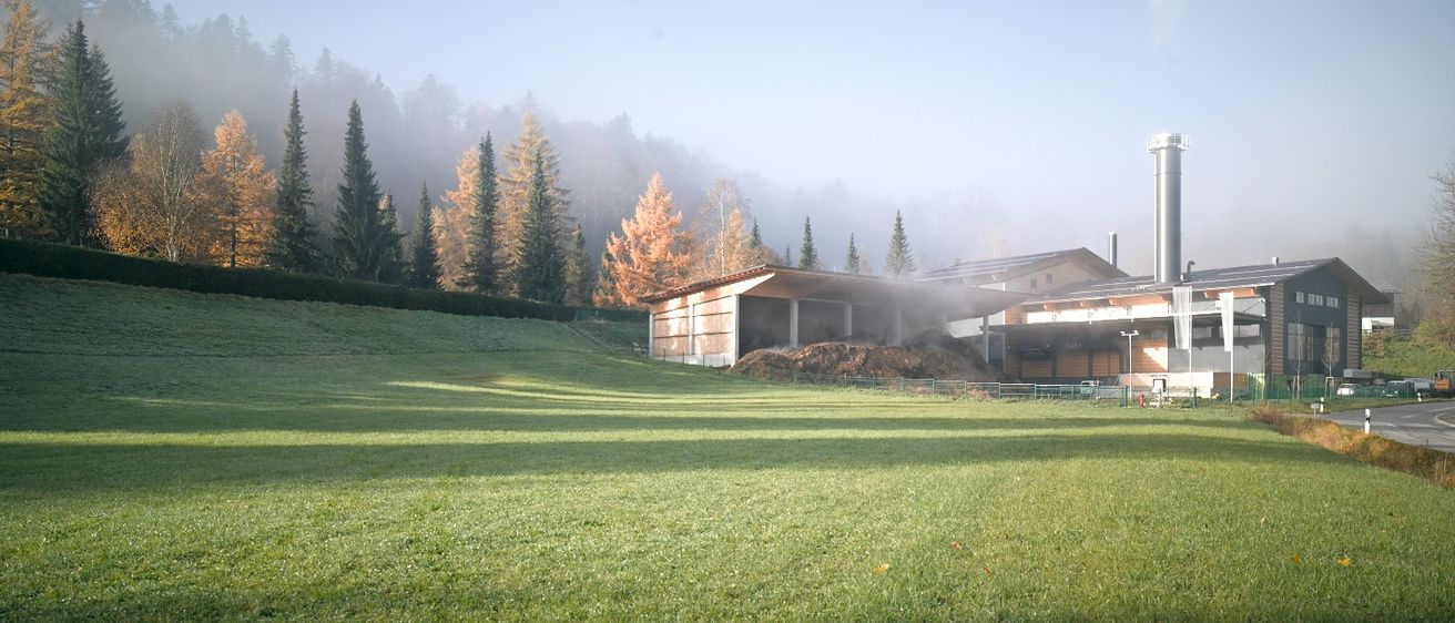 Ein Gebäude mit einem Schornstein stößt Rauch aus und befindet sich in einem weitläufigen Grasfeld mit Bäumen. Nebel umgibt die Bäume und der Himmel ist blau.