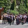 Ein Umzug von Männern und Frauen in traditioneller bayerischer Tracht, die in einer Reihe auf einer Straße marschieren. Sie spielen Instrumente, und einige halten Regenschirme.