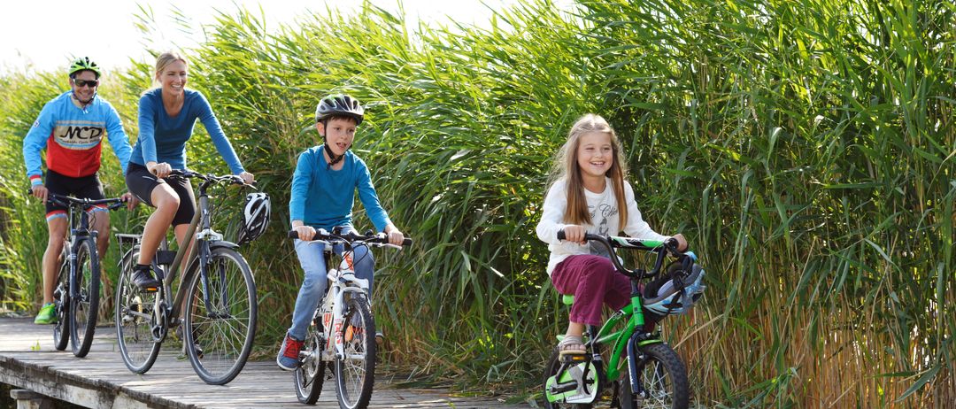 Drei Personen fahren auf einer Holzbrücke über Wasser mit Fahrrädern, umgeben von hohem Gras. Zwei Kinder lächeln, eines in Purpurrot und das andere in einem weißen Hemd.