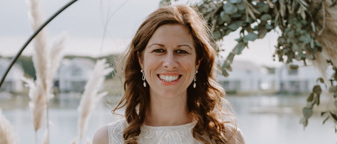 Eine lächelnde Frau in einem weißen Kleid steht am See und hält ein offenes Buch. Im Hintergrund befinden sich Grünpflanzen und ein See.